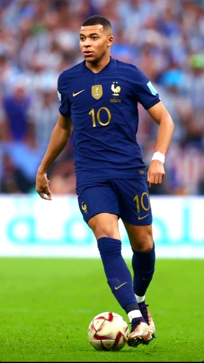 A male soccer player in a blue France national team uniform with the number 10 is dribbling a ball on the field during a match, as blurred spectators watch—perfect action to enjoy live on Latino IPTV.