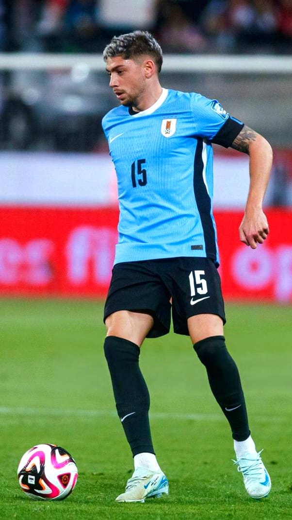 A soccer player in a light blue jersey with the number 15 controls the ball on the field during a match, perfect for watching live on Latino IPTV. He wears black shorts, black socks, and white shoes with a blurred stadium and red ad boards behind him.