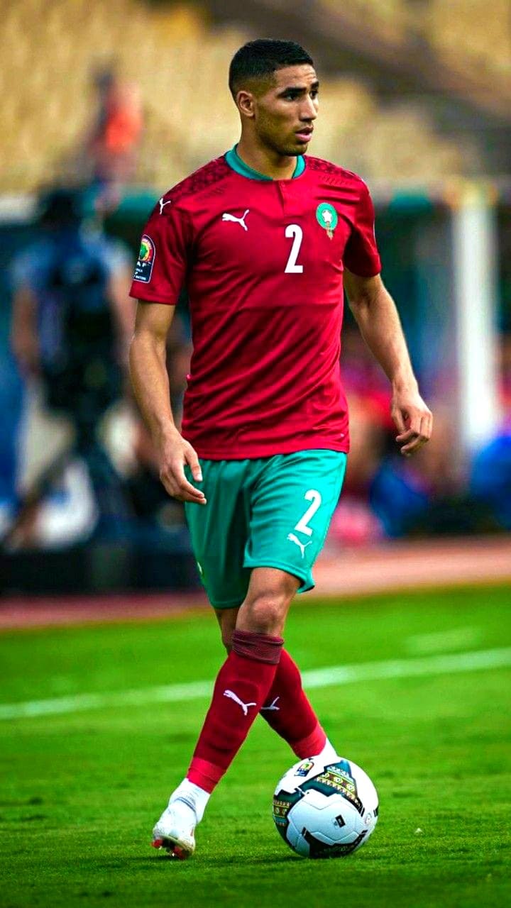 A male soccer player in a red and green uniform with the number 2 dribbles a soccer ball on a grassy field during a match, as fans enjoying the game through IPTV Latino watch from afar. The stadium and spectators are visible in the blurred background.